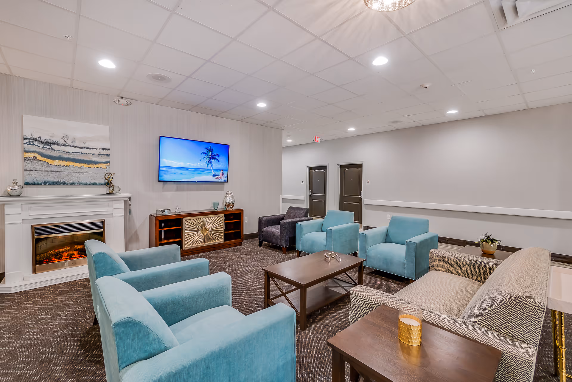 A cozy living room area in a senior living facility featuring a fireplace, a wall-mounted TV displaying a beach scene, four blue armchairs, one patterned sofa, a wooden coffee table, and a side table with a candle. The room has neutral-colored walls and carpeted flooring.