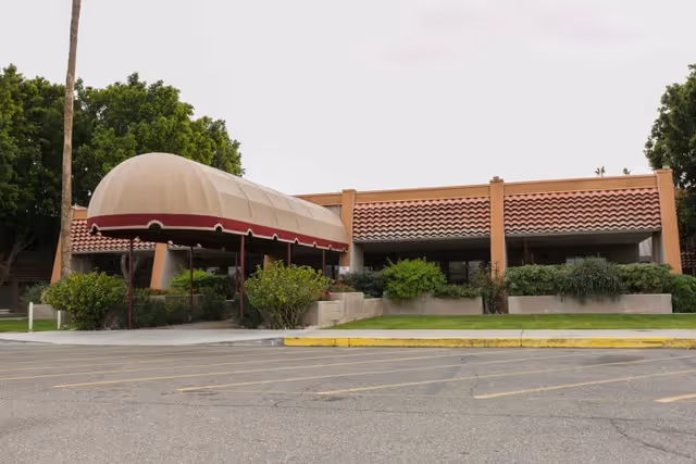 Front entrance of a single-story building with a tan-and-maroon covered awning, red tile roof, surrounding shrubs, and a parking lot.