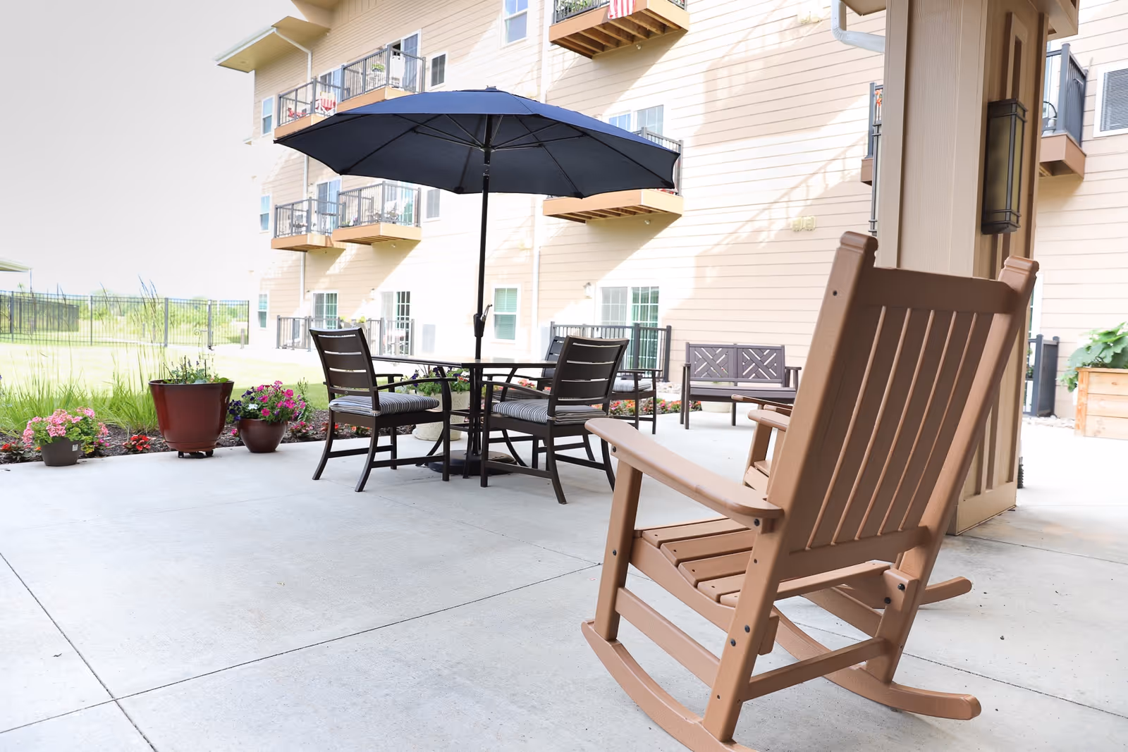 Outdoor patio area with a brown rocking chair in the foreground, a round table with four chairs and a large blue umbrella in the center, potted plants along the edge, and a multi-story building with balconies in the background.