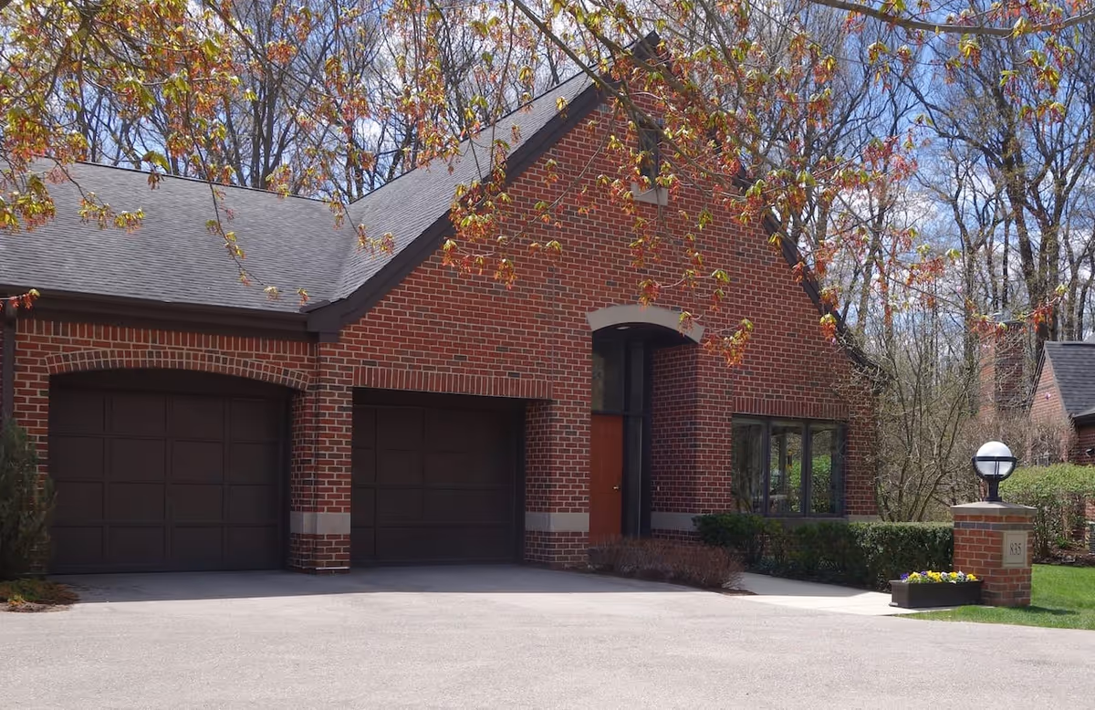 Exterior view of a brick building with two dark garage doors, a red entrance door, and a large window. There is a paved driveway in front, a lamp post on a brick pillar with the number 835, and some trees with budding leaves in the background.