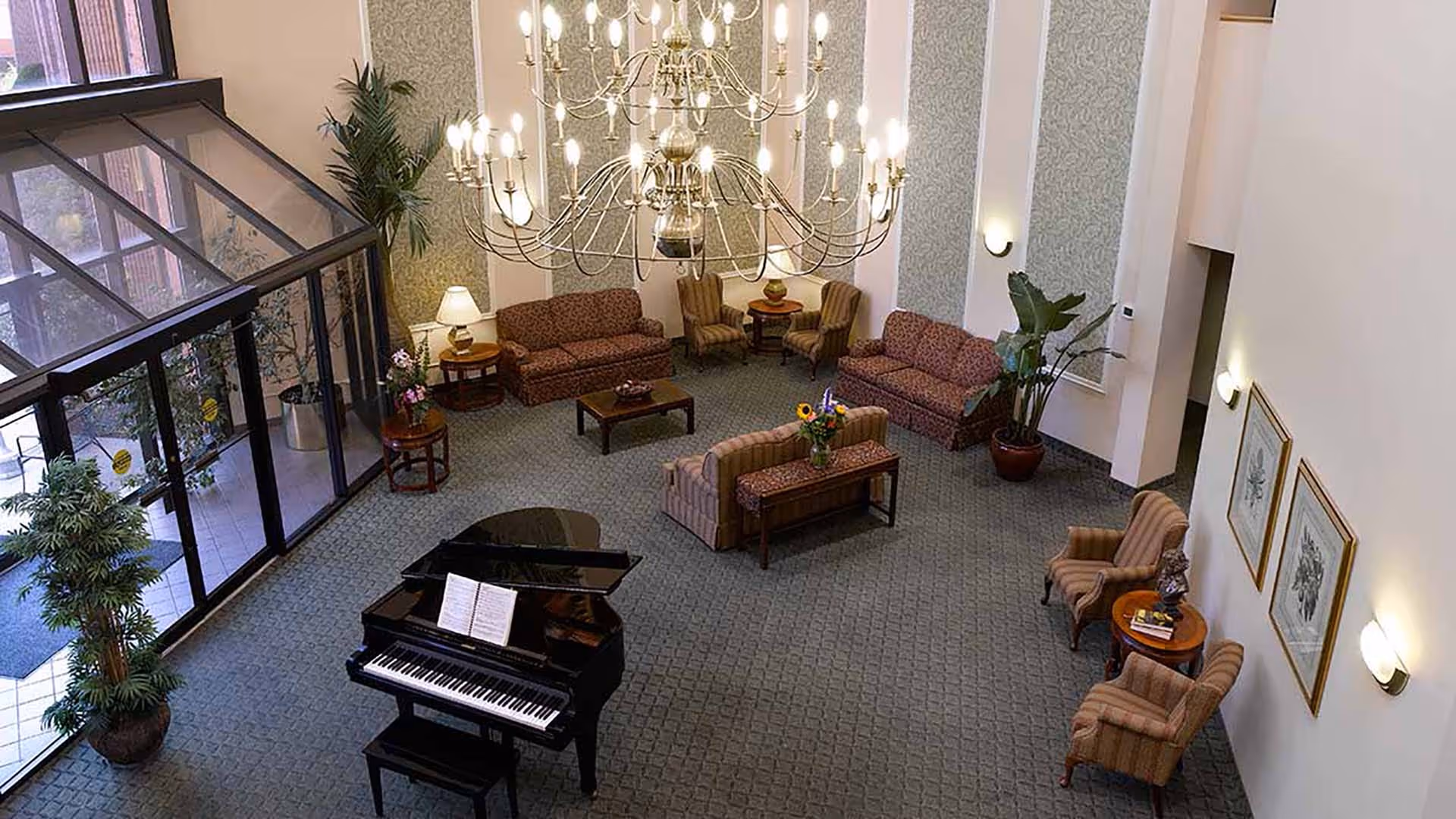 Spacious senior living community lounge area with a grand piano in the center, surrounded by multiple sofas and armchairs. Large chandelier hangs from the ceiling, and there are several potted plants and framed artwork on the walls. Glass doors lead to an outdoor area.