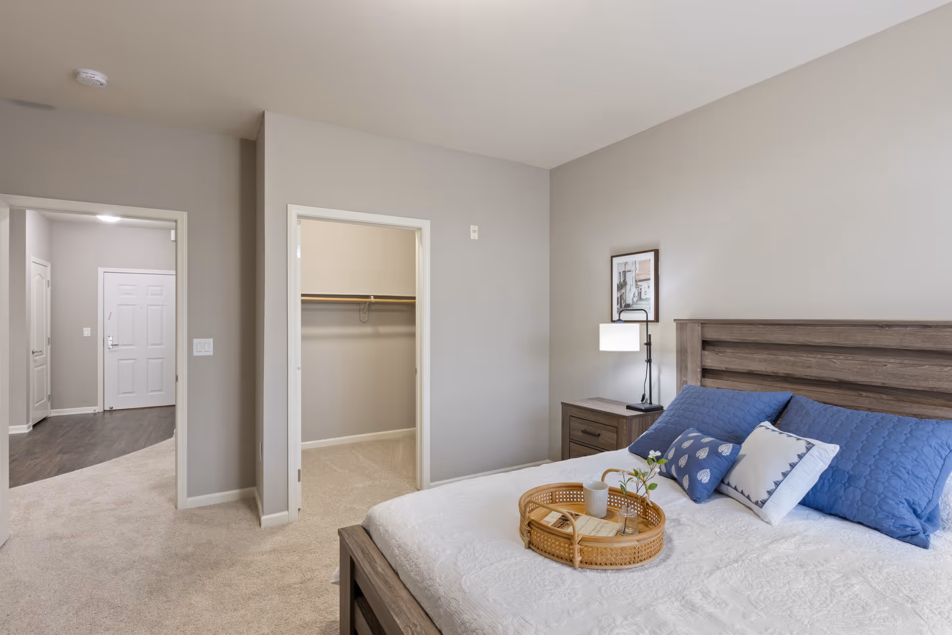 A bedroom in a senior living facility with a wooden bed frame, white bedspread, and blue pillows. A wicker tray with a cup, book, and small plant is on the bed. Next to the bed is a nightstand with a lamp and a framed picture on the wall. The room has beige carpet and light gray walls. An open doorway reveals a walk-in closet, and another doorway leads to a hallway with a white door at the end.