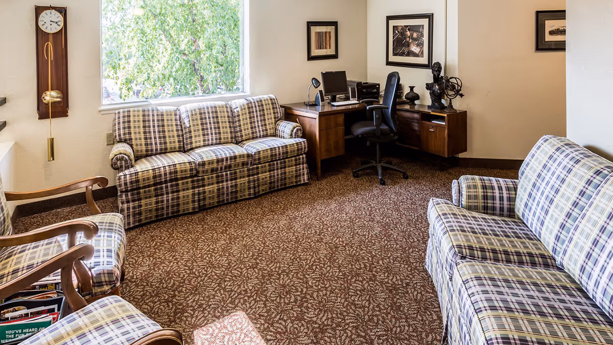 A cozy sitting area with plaid sofas and armchairs, a desk with an office chair, a wall clock, and a window showing trees outside.