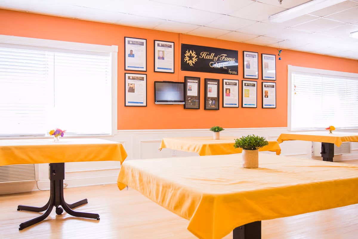 A bright dining room with orange walls and large windows covered with white blinds. Several tables are covered with yellow tablecloths and decorated with small green plants and colorful flowers. On the wall, there is a sign that reads 'Hall of Fame Cafe' surrounded by framed certificates and photos.