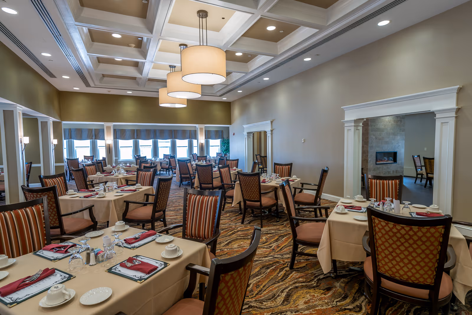 Spacious dining room with tables set for service, upholstered chairs, and large pendant lights under a coffered ceiling.