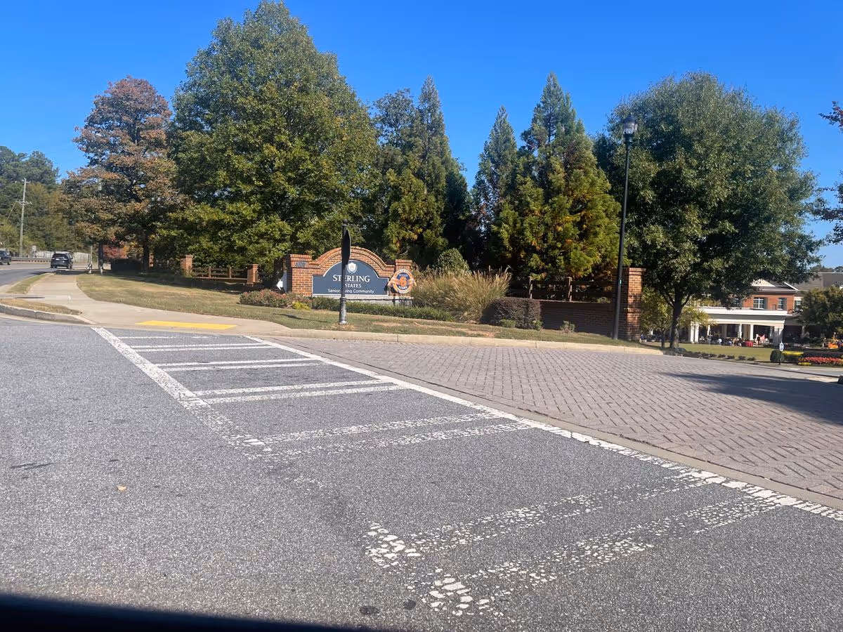 Entrance to Sterling Estates retirement community showing a brick sign, trees, and a paved road with a crosswalk in the foreground.
