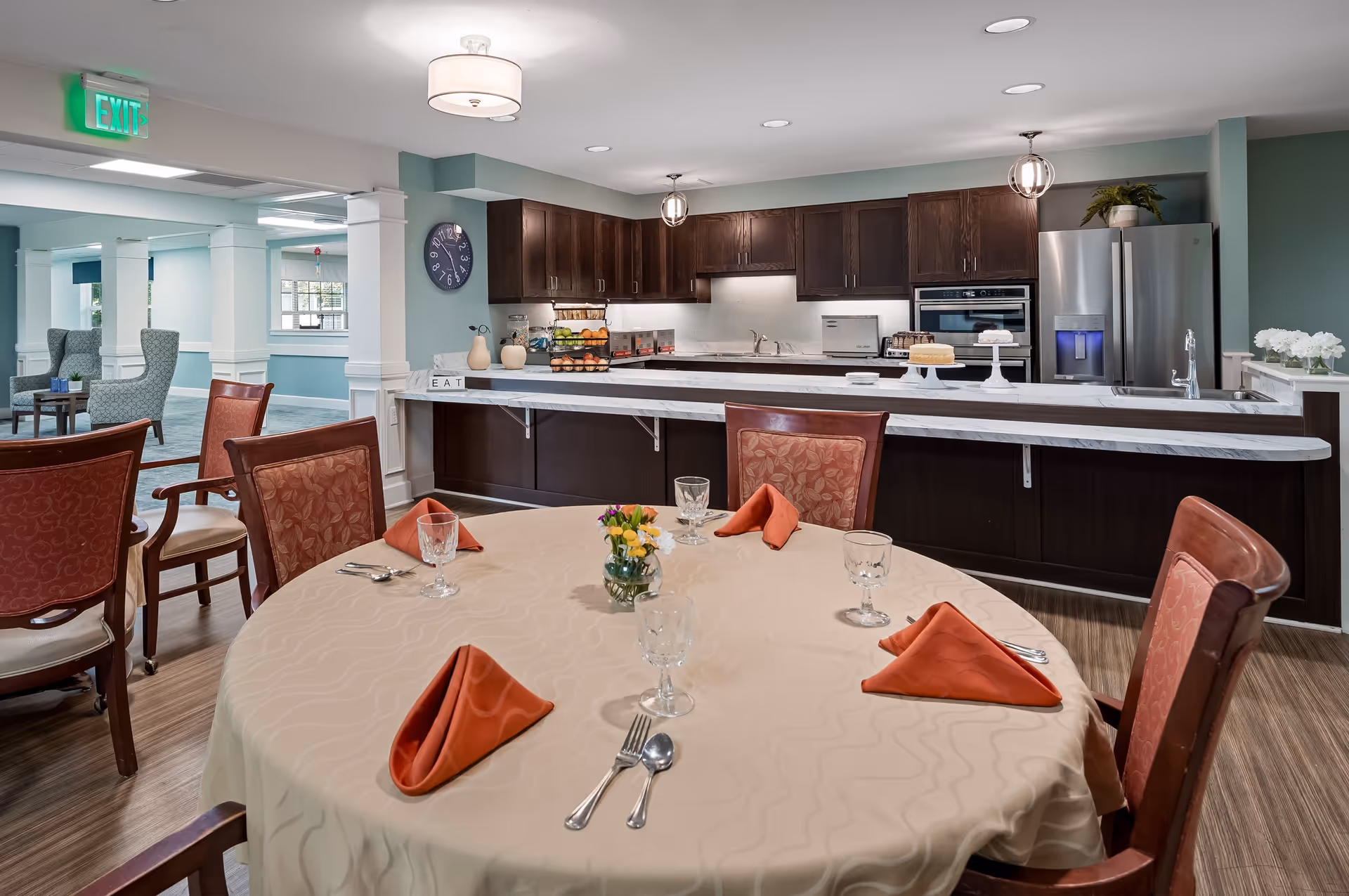 A dining area in a senior living facility featuring a round table set with a beige tablecloth, orange folded napkins, glassware, and silverware. In the background, there is a kitchen with dark wooden cabinets, a marble countertop, stainless steel refrigerator, oven, and various kitchen items. The room has soft lighting and a calm, inviting atmosphere with a seating area visible to the left.