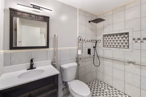 A modern bathroom with a white toilet, a dark wood vanity with a white countertop and black faucet, a large mirror with a dark wood frame, and a walk-in shower featuring a rainfall showerhead, handheld shower, and tiled walls with a decorative inset and grab bars.