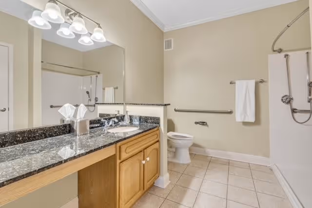 A spacious bathroom with beige walls and tiled floor. There is a large granite countertop with a sink, wooden cabinets underneath, and a large mirror above. The bathroom includes a toilet with grab bars on the wall and a walk-in shower with a handheld showerhead and a towel hanging on a bar.