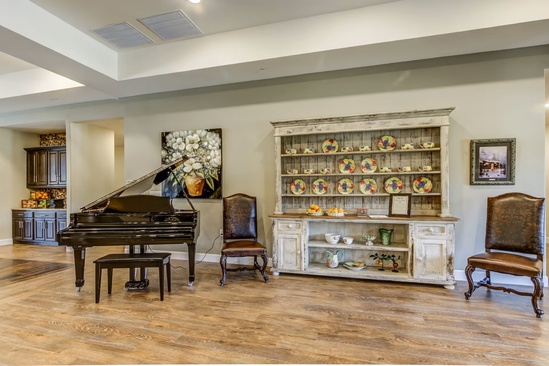 A spacious room with a black grand piano and bench on the left, two ornate wooden chairs with leather seats on either side of a rustic white wooden hutch displaying decorative plates and ceramics. The room has wooden flooring, light-colored walls, and framed artwork hanging on the walls.