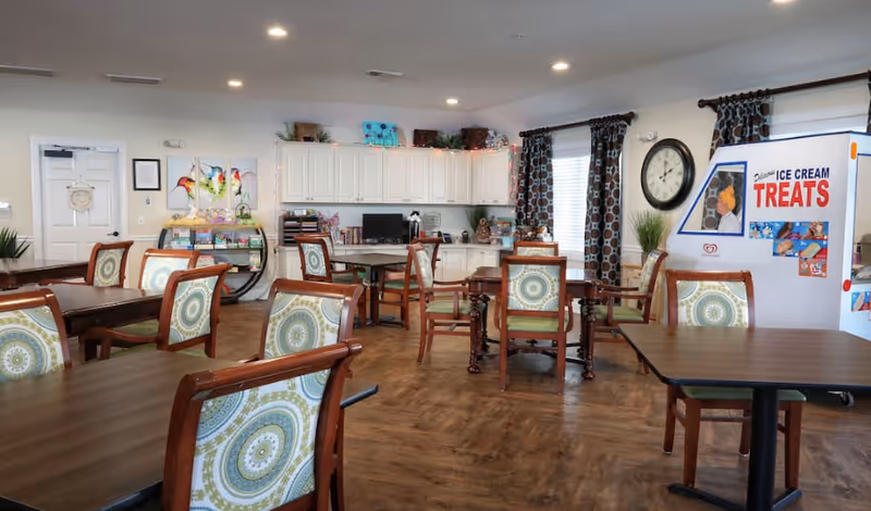 A bright and spacious common area with multiple wooden tables and chairs featuring patterned cushions. The room has wood flooring, white cabinets along the back wall, a computer on a desk, colorful bird artwork on the wall, and a large clock. There is an ice cream treats vending machine on the right side near windows with patterned curtains.
