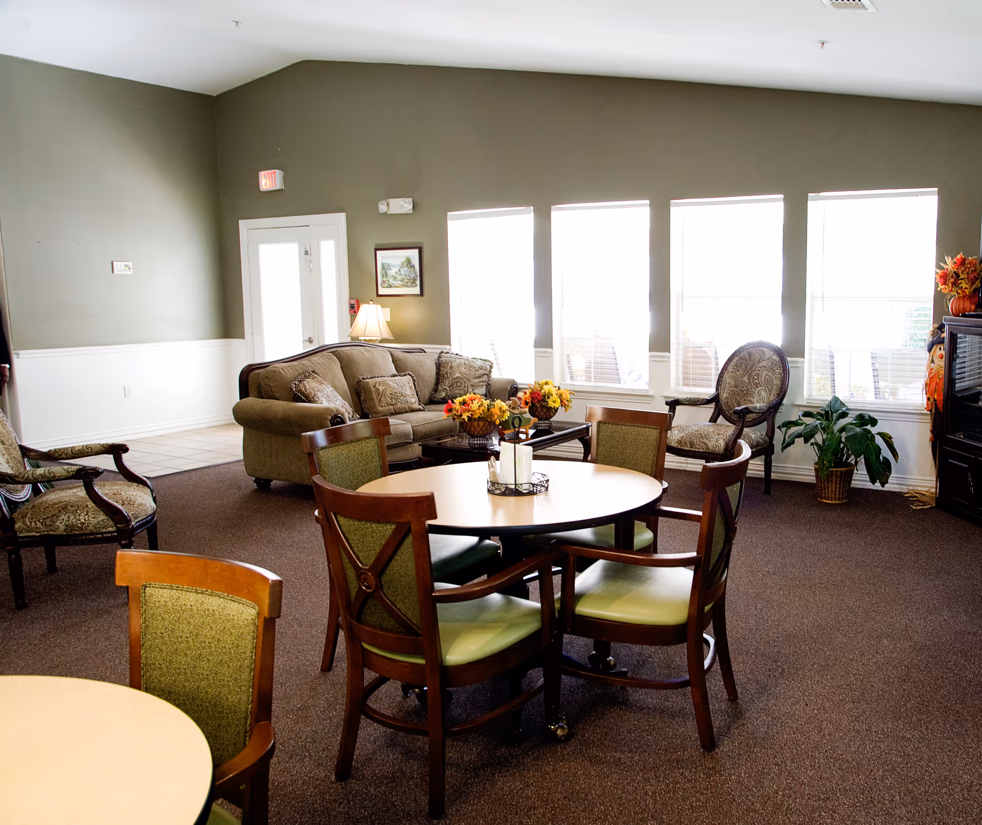 Communal living room with round tables and chairs, a sofa and armchairs arranged near large windows.