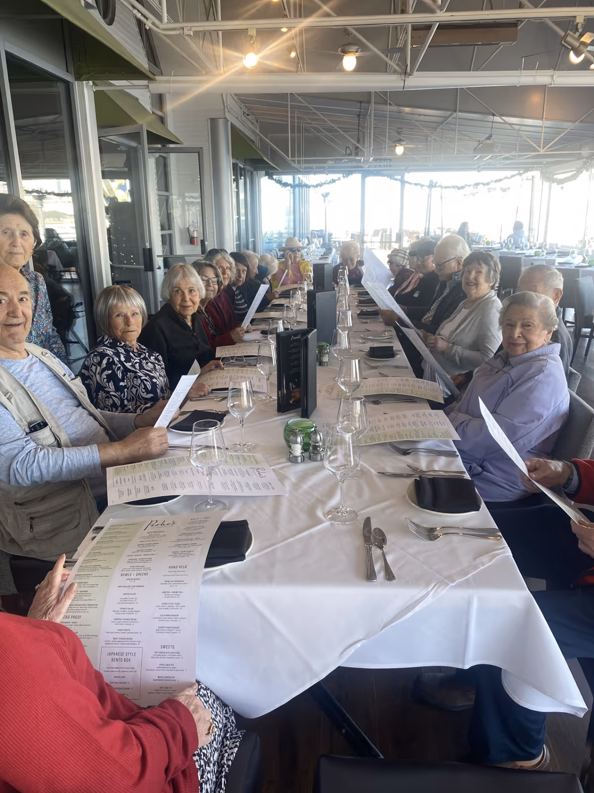 A group of elderly people sitting around a long dining table in a bright restaurant, holding menus and preparing to order food. The table is set with white tablecloths, wine glasses, and silverware. The background shows large windows with a view outside.