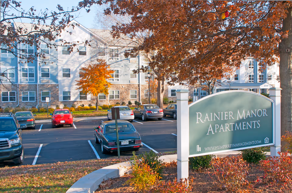 Exterior view of Rainier Manor Apartments with a parking lot in front. Several cars are parked and autumn trees with orange leaves surround the area. The apartment building is multi-story with white and stone facade.