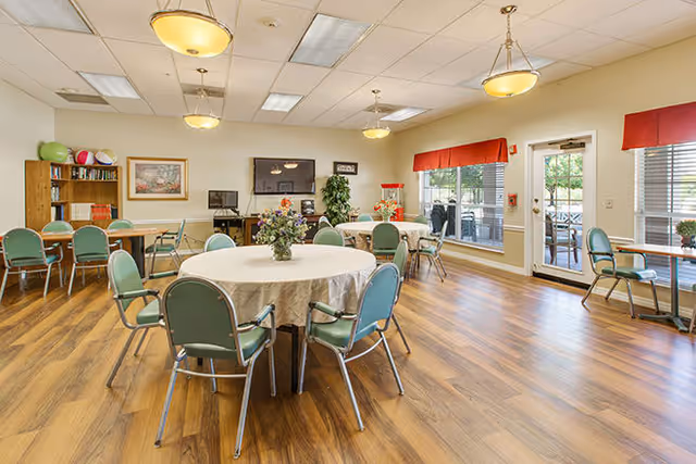 A bright and spacious common area with round tables covered in white tablecloths, surrounded by green chairs. The room has wood flooring, large windows with red valances, and glass doors leading outside. There is a TV mounted on the wall, a bookshelf with books and decorative items, and ceiling lights providing illumination.