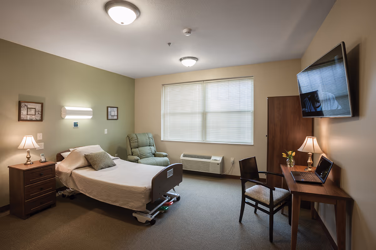A furnished resident bedroom with an adjustable bed, nightstand, recliner, desk and wall-mounted TV.