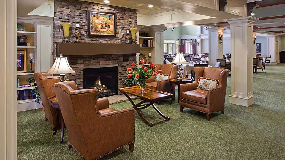 A cozy seating area with four brown leather armchairs arranged around a wooden coffee table with a vase of red flowers, set in front of a stone fireplace with a painting above it. The room has green carpet and is well-lit with table lamps. In the background, there is a dining area with tables and chairs.
