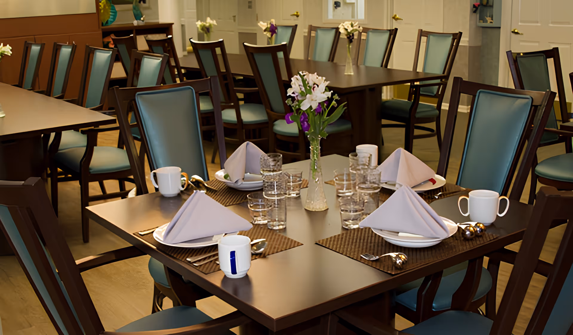 Dining room with multiple tables set with folded napkins, cups, glasses, and vases of flowers.