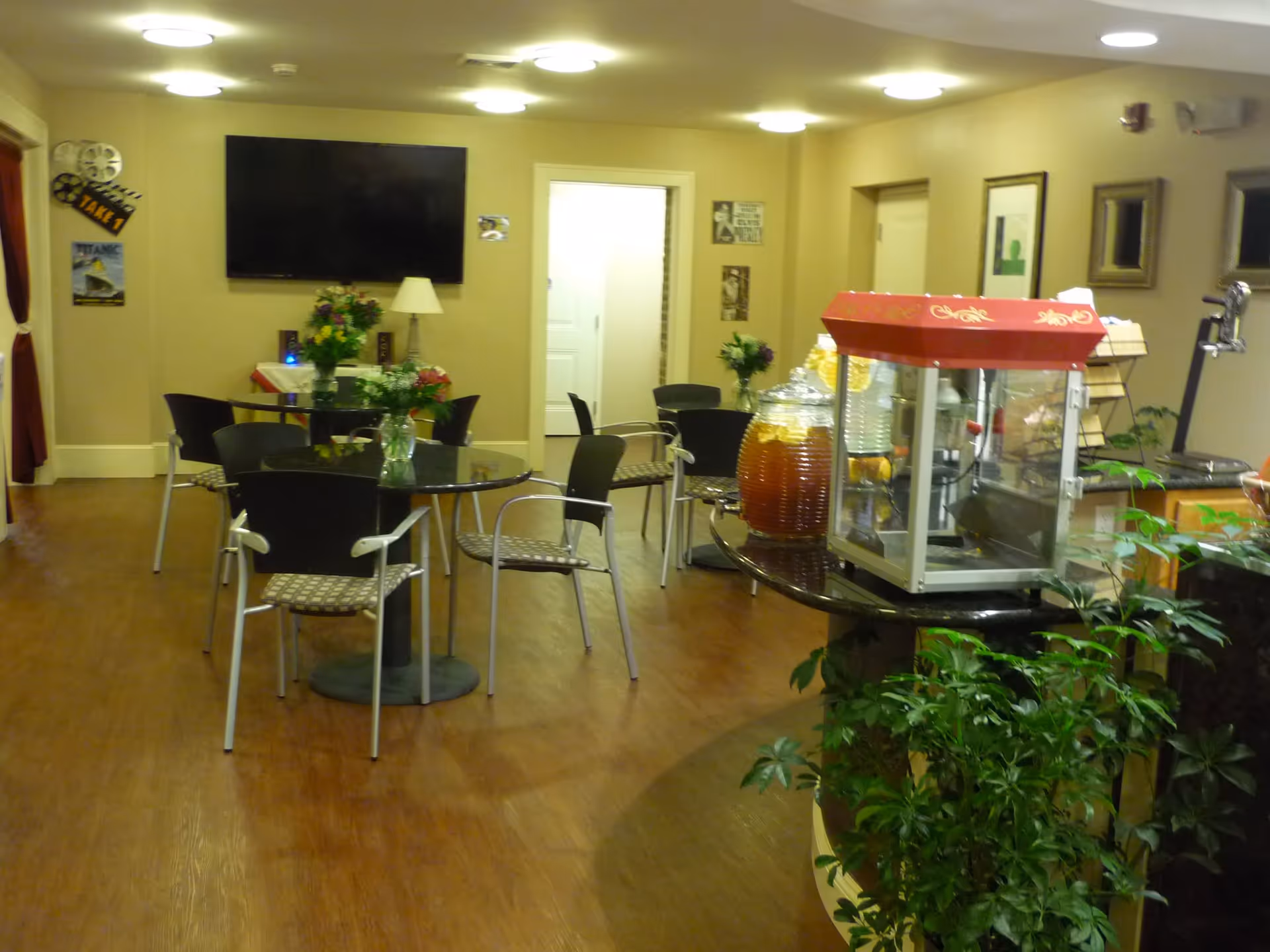Interior view of a senior community common area with round tables and chairs arranged for seating. A large flat-screen TV is mounted on the wall, and there are decorative flowers on the tables. A popcorn machine and beverage dispensers are on a counter to the right, with some green plants nearby. The room has warm lighting and framed pictures on the walls.