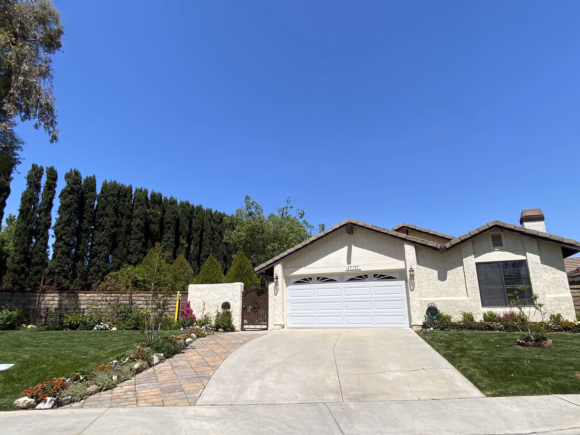 Single-story house with a white garage door, beige stucco exterior, and a brown tiled roof. The driveway is concrete with a brick-paved walkway on the left side leading to a wooden gate. The front yard has green grass, flower beds, and tall trees along the back fence under a clear blue sky.