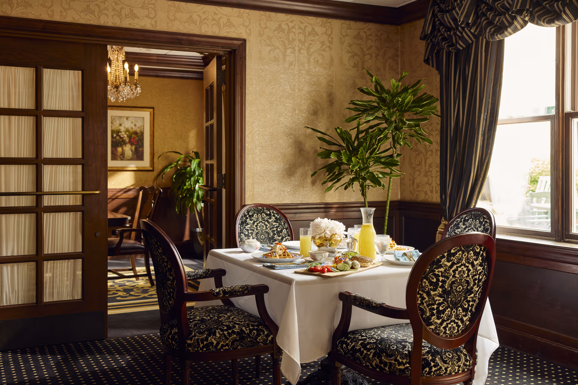 A dining room with a table set for breakfast, featuring plates of food, glasses of juice, and a vase with white flowers. The room has ornate wallpaper, dark wood paneling, patterned upholstered chairs, and a large window with striped curtains letting in natural light. A doorway with glass-paneled wooden doors leads to another room with a chandelier and framed artwork.