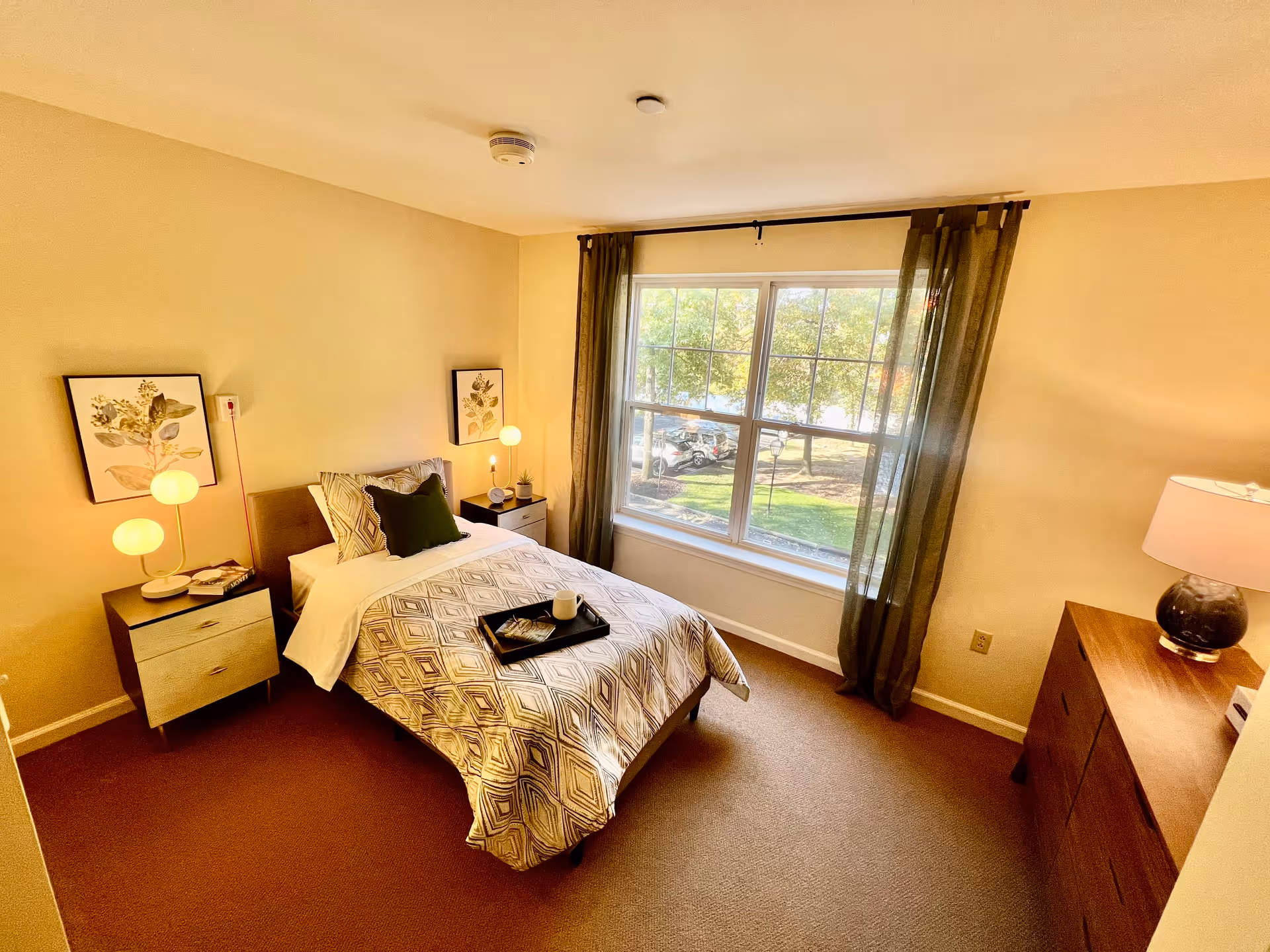 A cozy bedroom with a single bed covered in a patterned duvet and pillows, two bedside tables with lamps and decorative items, a large window with sheer curtains letting in natural light, and a wooden dresser with a lamp on top.