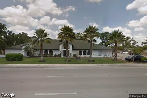 Single-story building with a green roof and white exterior walls, surrounded by palm trees and a well-maintained lawn, viewed from across a street under a partly cloudy sky.