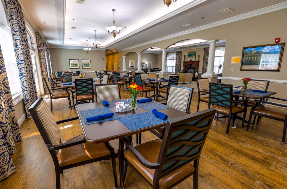 Spacious dining room with multiple set tables and chairs, chandeliers, large windows with patterned curtains, and floral centerpieces.