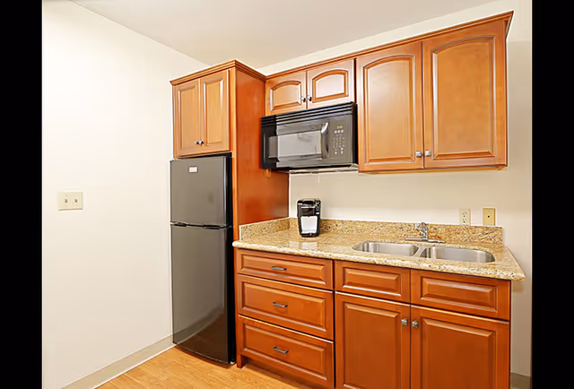 Small kitchenette with wooden cabinets, granite countertop, double sink, microwave, mini refrigerator, and a coffee maker.