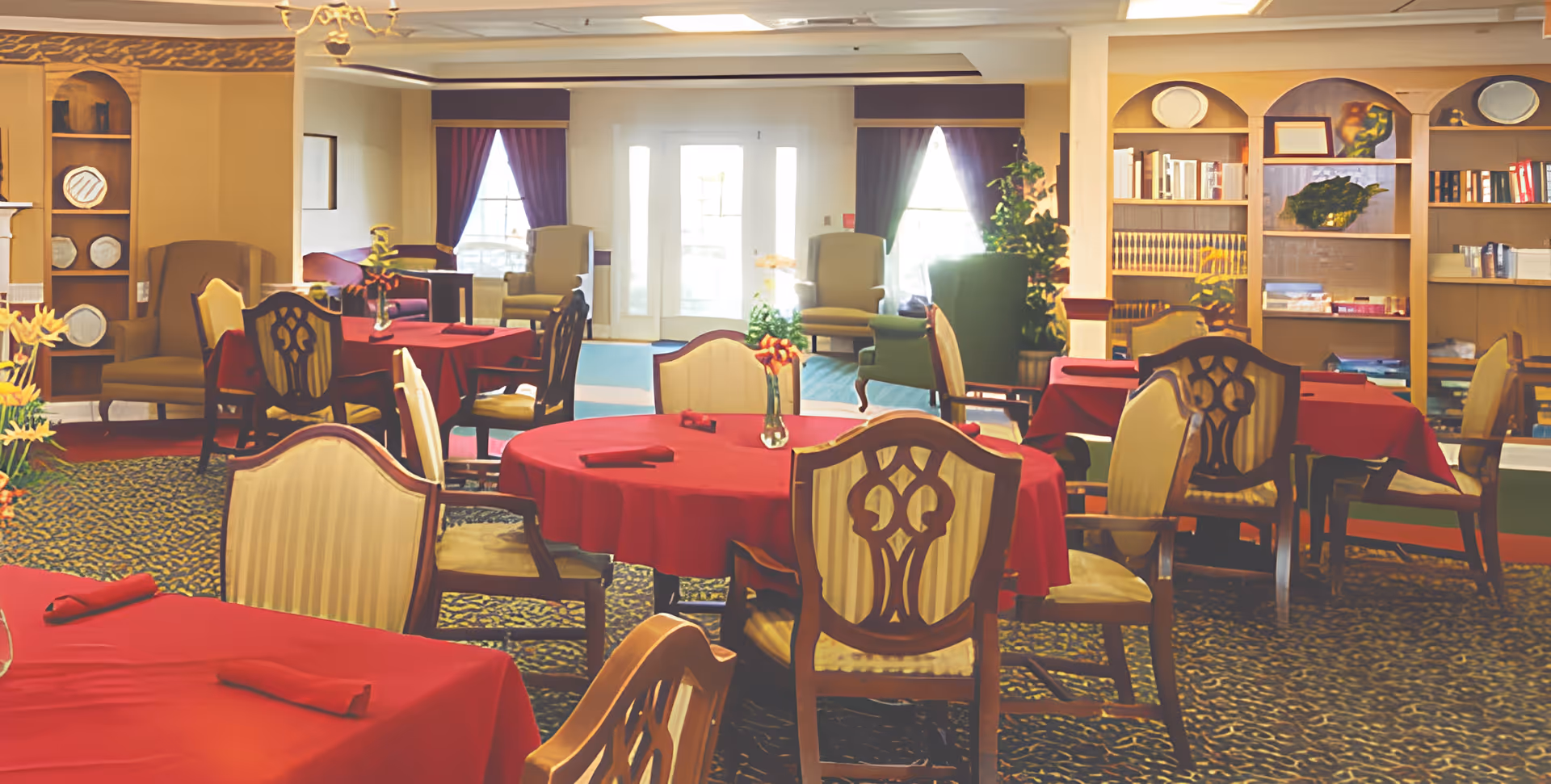 A dining room with several round tables covered in red tablecloths and matching red napkins. The chairs have wooden frames with cushioned seats and backs. The room features carpeted flooring with a patterned design, built-in wooden shelves with books and decorative plates, and large windows with dark curtains allowing natural light to enter. There are also a few armchairs near the windows and some plants adding greenery to the space.
