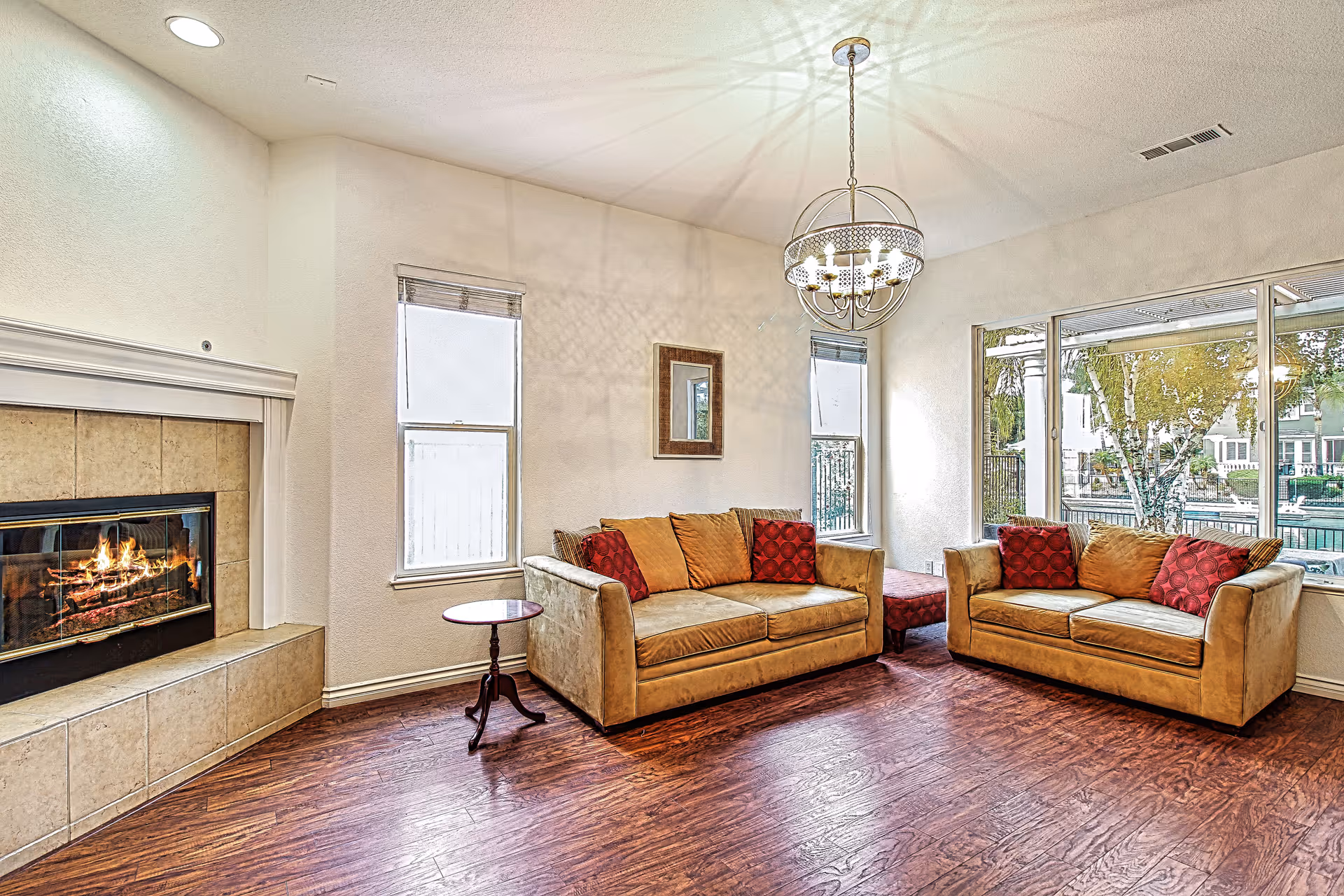 A cozy living room with two beige sofas adorned with red and beige cushions, a small round wooden side table, a fireplace with a burning fire on the left, wooden flooring, two tall windows, a large window showing an outdoor view, and a decorative chandelier hanging from the ceiling.