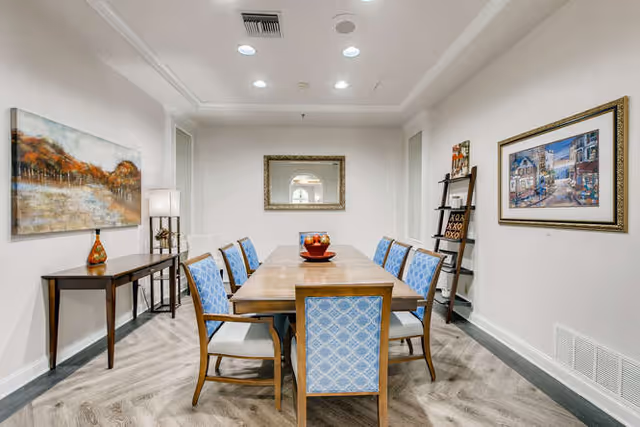 A dining room with a wooden table surrounded by eight chairs with blue patterned upholstery. The room has light-colored walls and wood flooring. On the left wall, there is a large painting above a narrow wooden console table with a decorative vase and lamp. On the right wall, there is a framed picture and a leaning ladder shelf with decorative items. A rectangular mirror is centered on the far wall above the table.