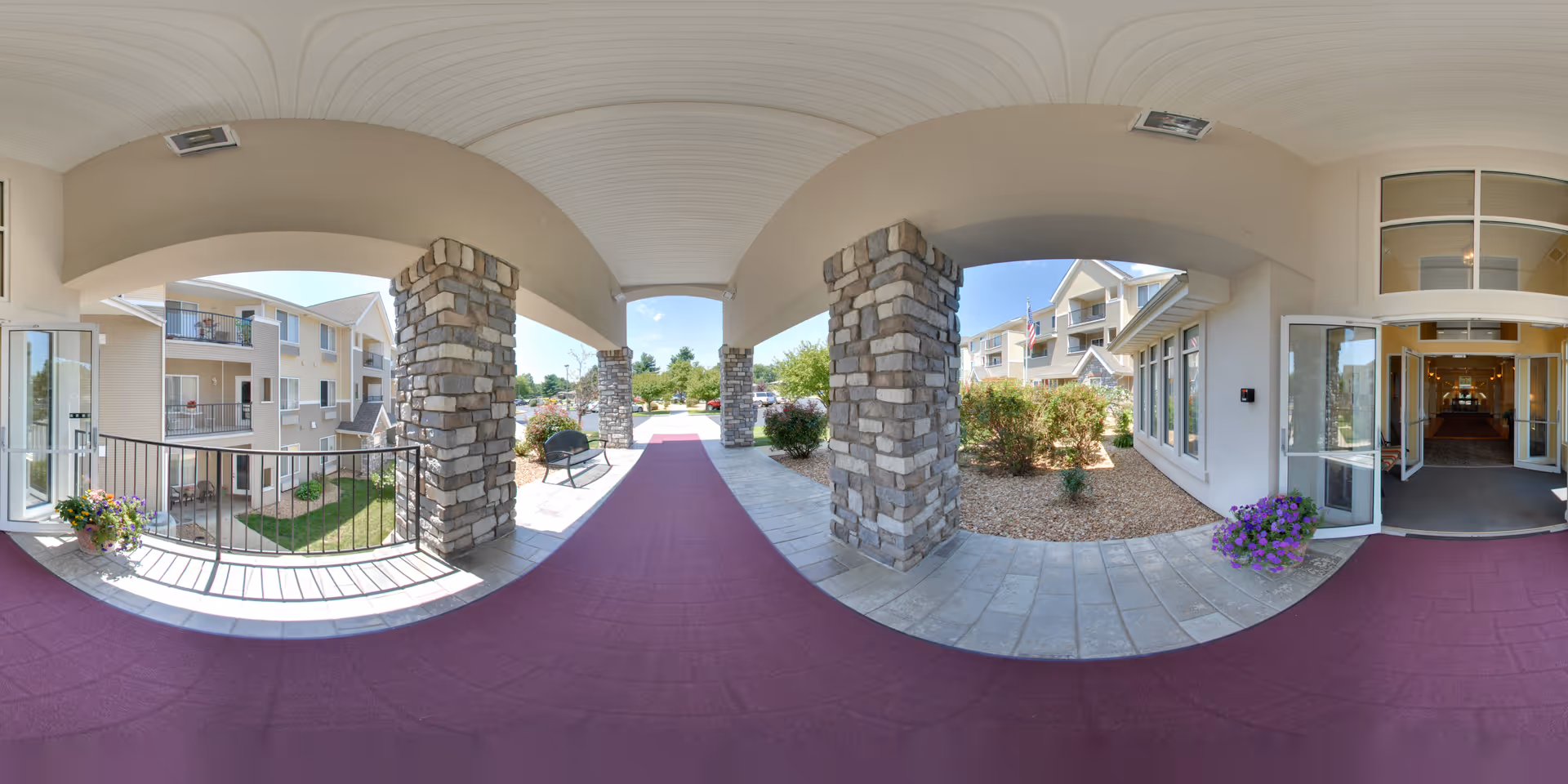 Covered entrance area of College View Manor with stone pillars, a maroon carpet runner, potted flowers, and a bench. The entrance leads into a building with glass doors and windows, and the exterior shows a multi-story residential building with balconies and landscaping.