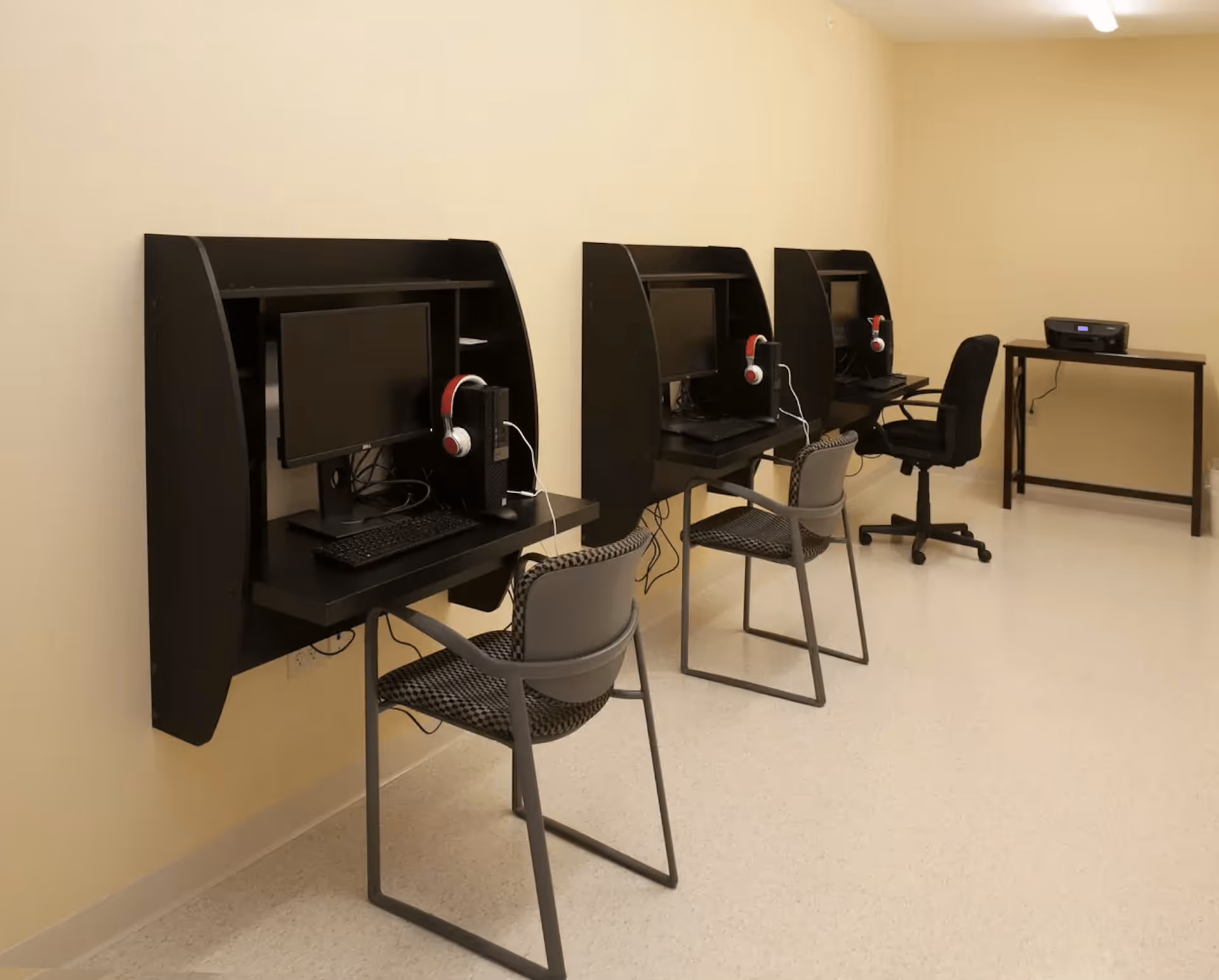 A room with three black computer workstations mounted on a beige wall, each equipped with a monitor, keyboard, desktop computer, and red and white headphones. Each workstation has a chair in front of it. There is also a small table with a printer on it against the far wall.