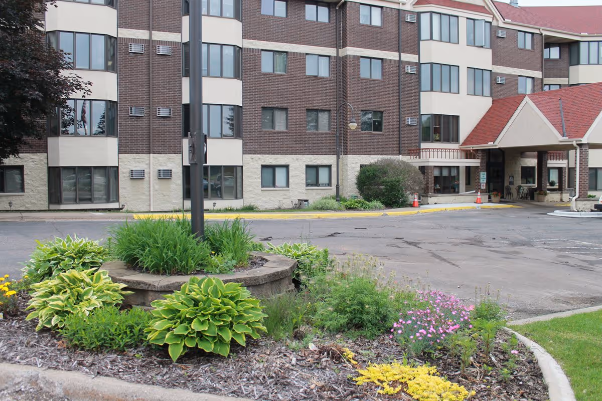 Exterior view of Gracepointe Crossing Gables, showing a multi-story brick and beige building with multiple windows, a red-roofed entrance canopy, a parking area, and landscaped garden beds with green plants and flowers in the foreground.