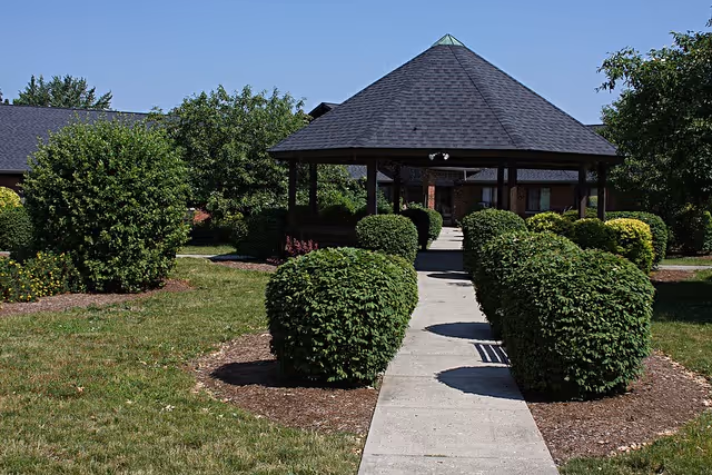 A paved walkway lined with neatly trimmed bushes leads to a covered gazebo structure with a dark shingled roof. The surrounding area features green grass, additional bushes, and trees under a clear blue sky.