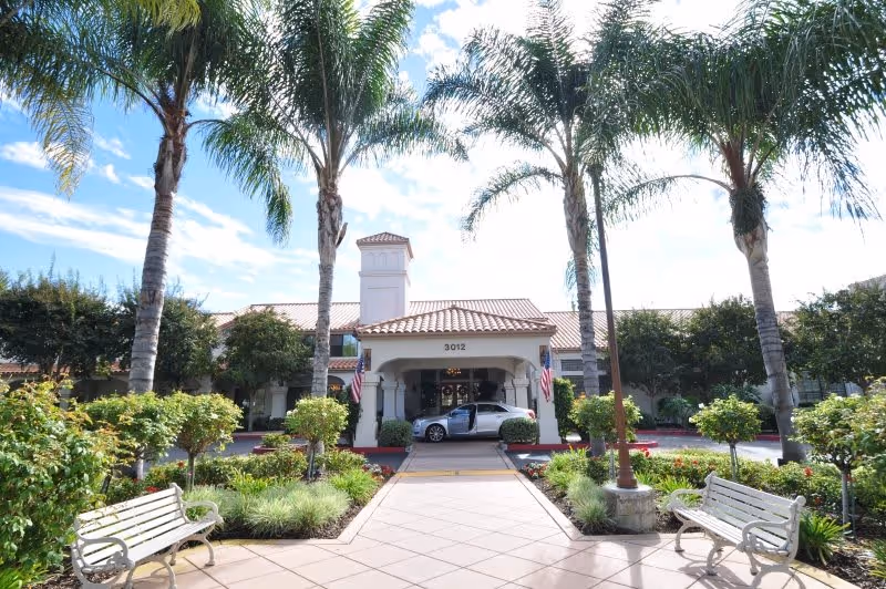 Front exterior view of Oakmont of Escondido Hills facility with a tiled roof entrance, palm trees, benches, and a car parked under the covered driveway.