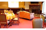 A cozy common area with a round dining table set with plates and cups, surrounded by chairs. There is a patterned sofa and armchair near a brick fireplace. The room has warm lighting and carpeted flooring.