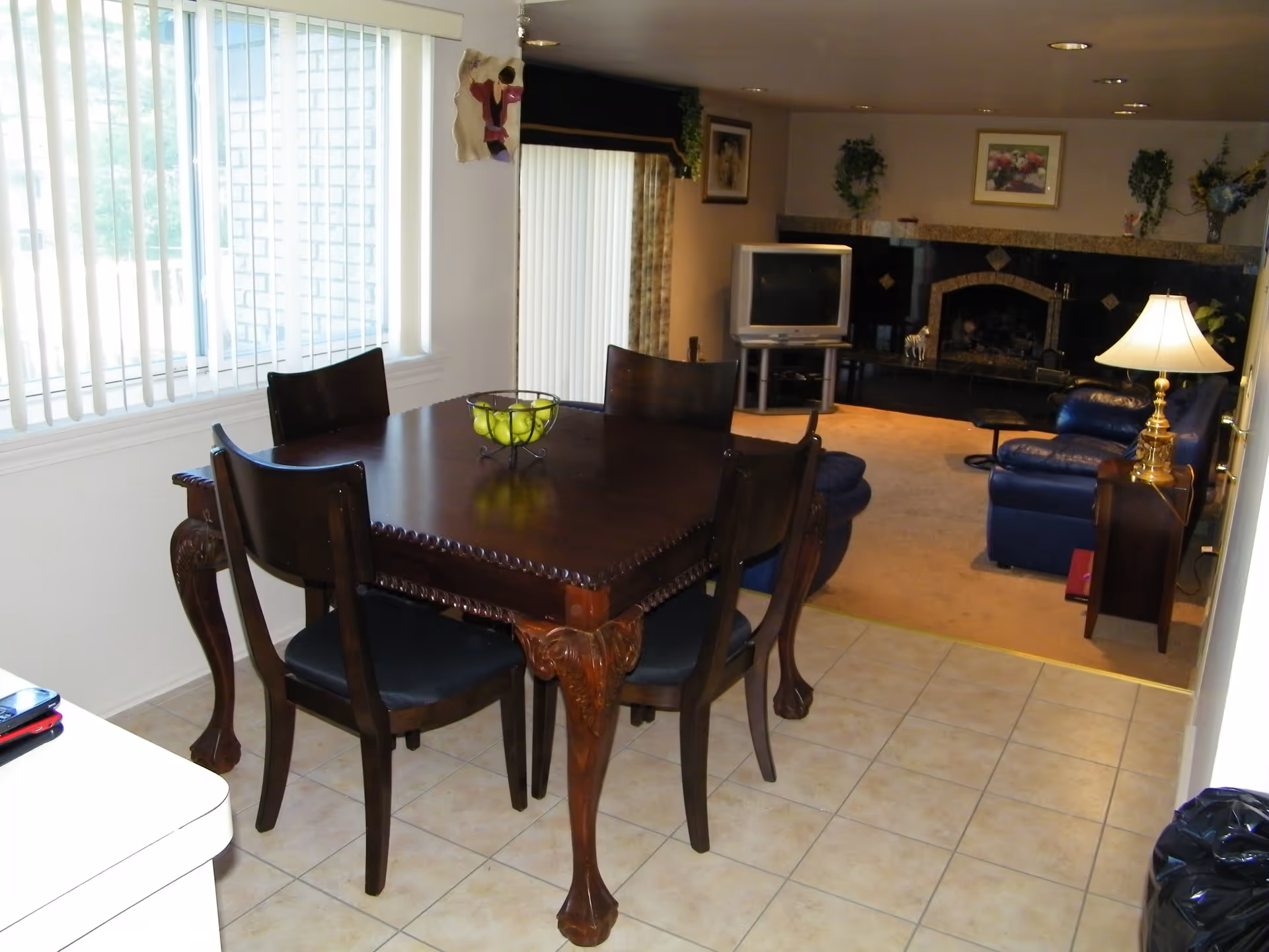 A dining area with a dark wooden table and four matching chairs on a tiled floor. A bowl of green apples is on the table. In the background, there is a living room with blue leather chairs, a TV on a stand, a fireplace with decorative tiles, plants, and framed pictures on the walls. A lamp on a side table is also visible.