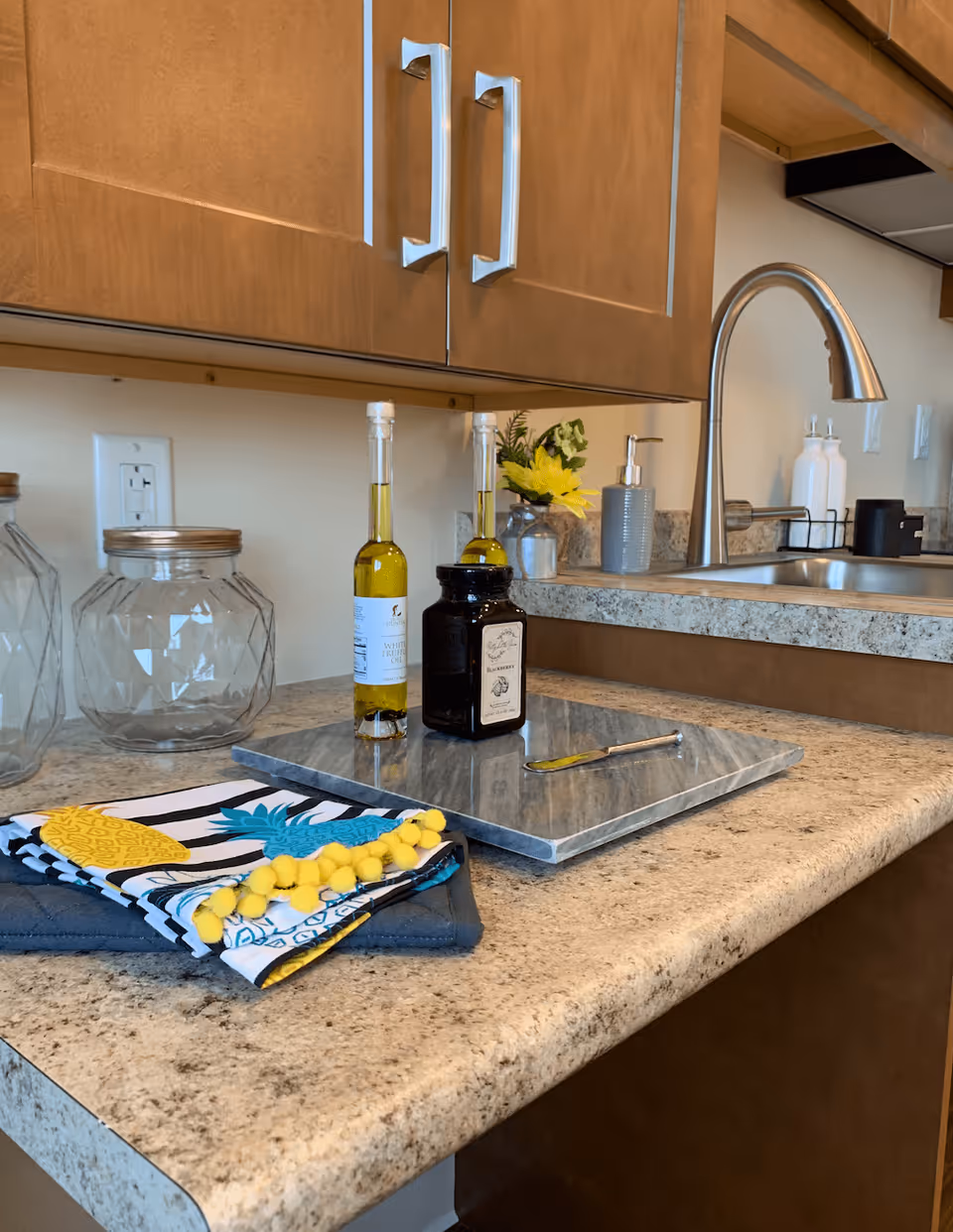 A kitchen countertop with a granite-like surface featuring a cutting board, two bottles of white truffle oil, a jar labeled blackberry, a knife, and colorful kitchen towels with pineapple designs. Above the counter are wooden cabinets with silver handles, and a stainless steel faucet and sink are visible in the background. A small flower arrangement and soap dispenser are also on the counter near the sink.