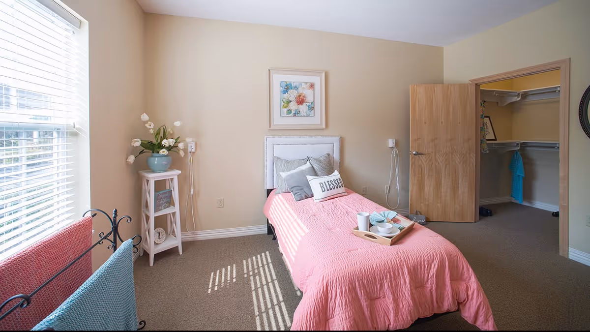 A bright bedroom with a single bed covered in a pink quilt and several pillows, including one with the word 'BLESSED'. There is a small white side table with a vase of flowers next to the bed, a window with blinds letting in natural light, and an open door leading to a walk-in closet with hanging clothes.