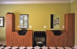 Interior view of a salon area in a senior living facility with two wooden cabinets and black sinks with mirrors above them, set against a light green wall. Between the cabinets is a black cushioned bench with magazines underneath. The floor has a black and white checkered pattern.