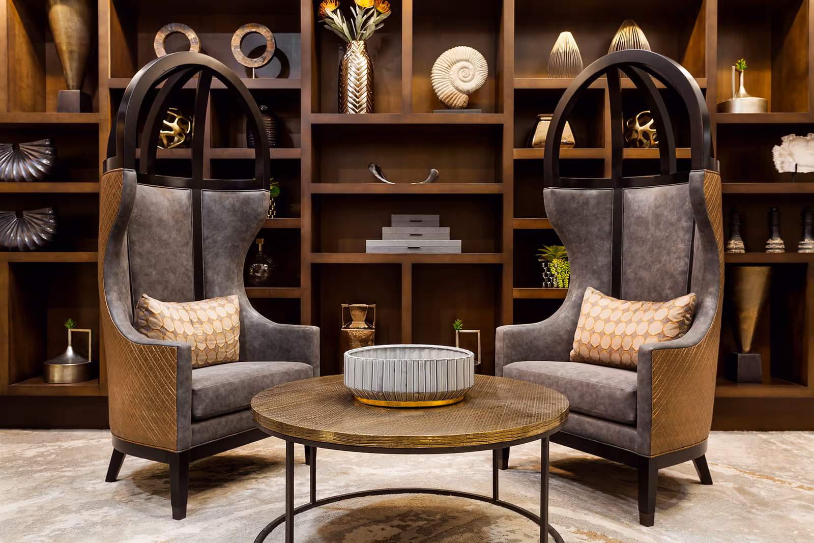 Two high-backed, modern armchairs with patterned cushions facing each other across a round wooden coffee table with a decorative bowl on top, set in front of a large wooden shelving unit filled with various decorative objects and plants.