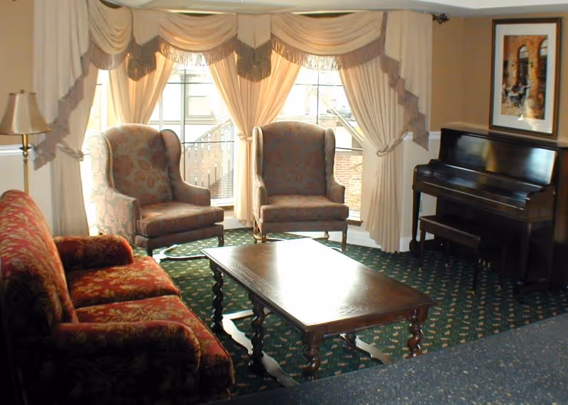 A cozy living room with a patterned red sofa, two upholstered armchairs, a wooden coffee table, and a dark wood upright piano. The room has large windows with cream-colored drapes and valances, and a framed picture hangs on the wall above the piano.