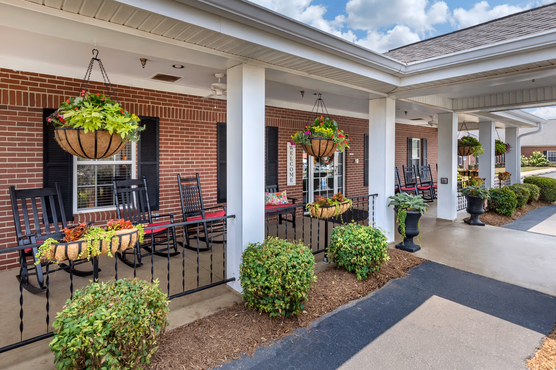 Covered outdoor porch area at Wesley Court Assisted Living Community with hanging flower baskets, black rocking chairs with red cushions, potted plants, and a brick wall with windows and a door that has a welcome sign.