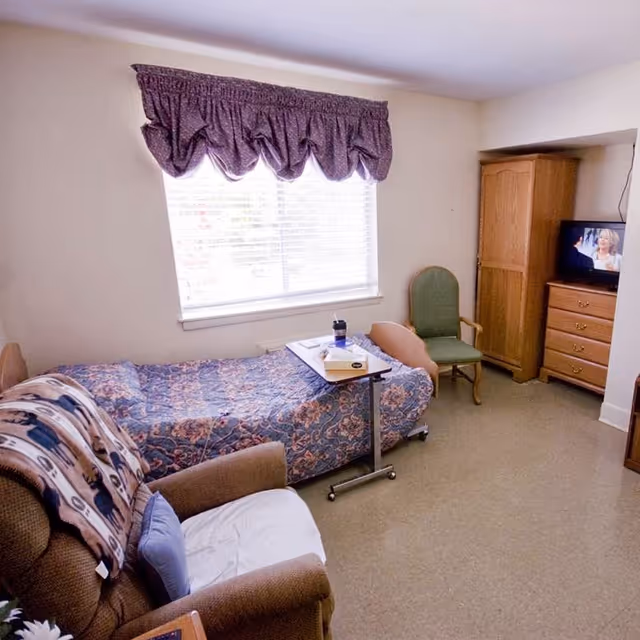 A small, cozy bedroom in a senior living facility with a single bed covered in a floral quilt, a green armchair, a wooden wardrobe, a chest of drawers with a small TV on top, and a recliner chair with a patterned blanket and a blue pillow. A window with blinds and a purple valance lets in natural light.