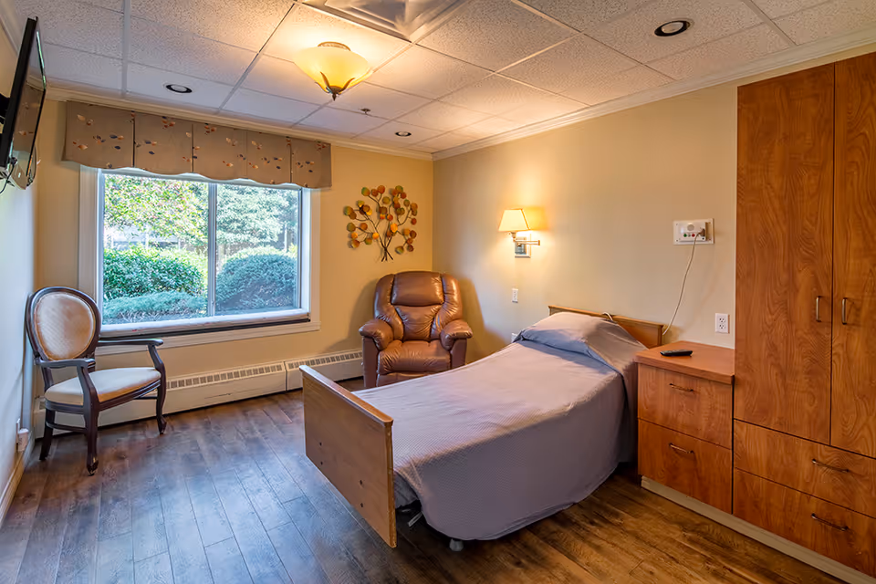 A bright private senior bedroom featuring a single bed, leather recliner, wooden cabinetry, a chair, and a large window overlooking landscaping.