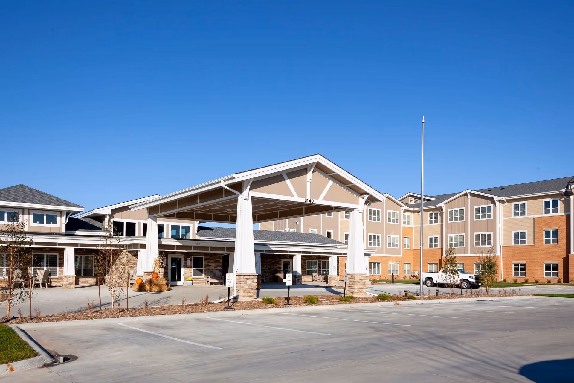 Exterior view of Cedarhurst Senior Living of La Vista building under a clear blue sky, featuring a covered entrance with stone pillars, multiple windows, and a parking area in front.