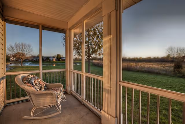 A cozy screened-in porch with a cushioned wicker rocking chair overlooking a grassy field and trees under a clear sky during sunset.
