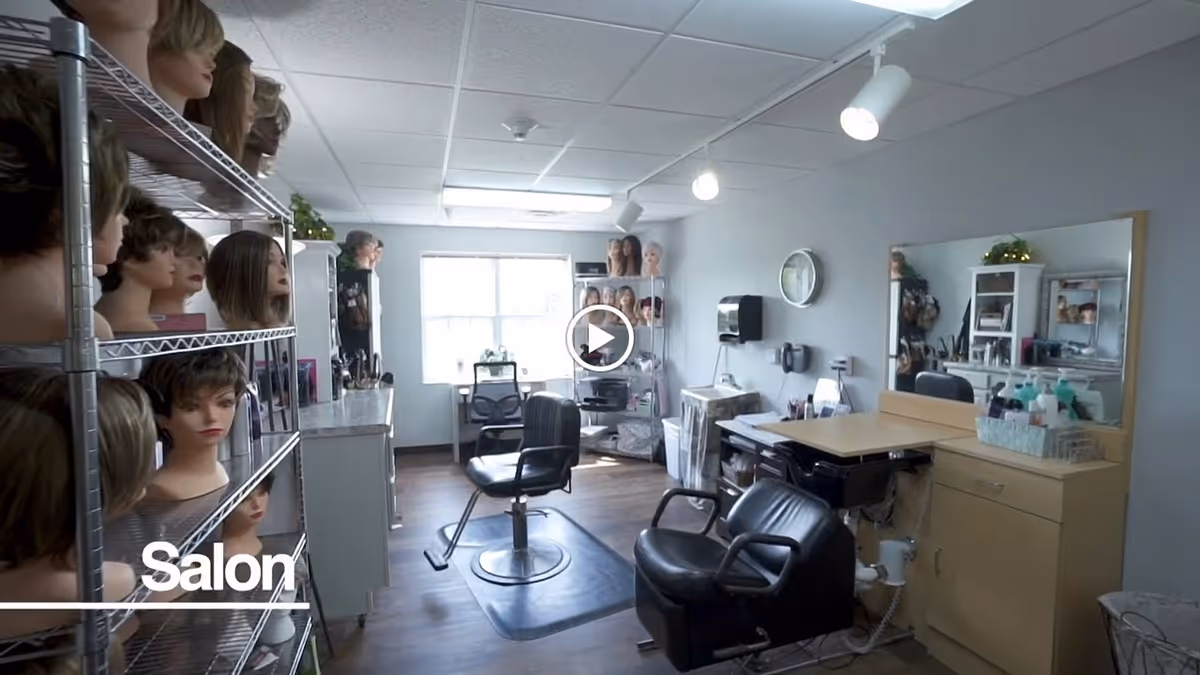 Interior view of a salon room with multiple mannequin heads displaying wigs on metal shelves to the left. The room has salon chairs, a large mirror, and various hair care products on a counter. The floor is wooden, and there is a window letting in natural light at the back.